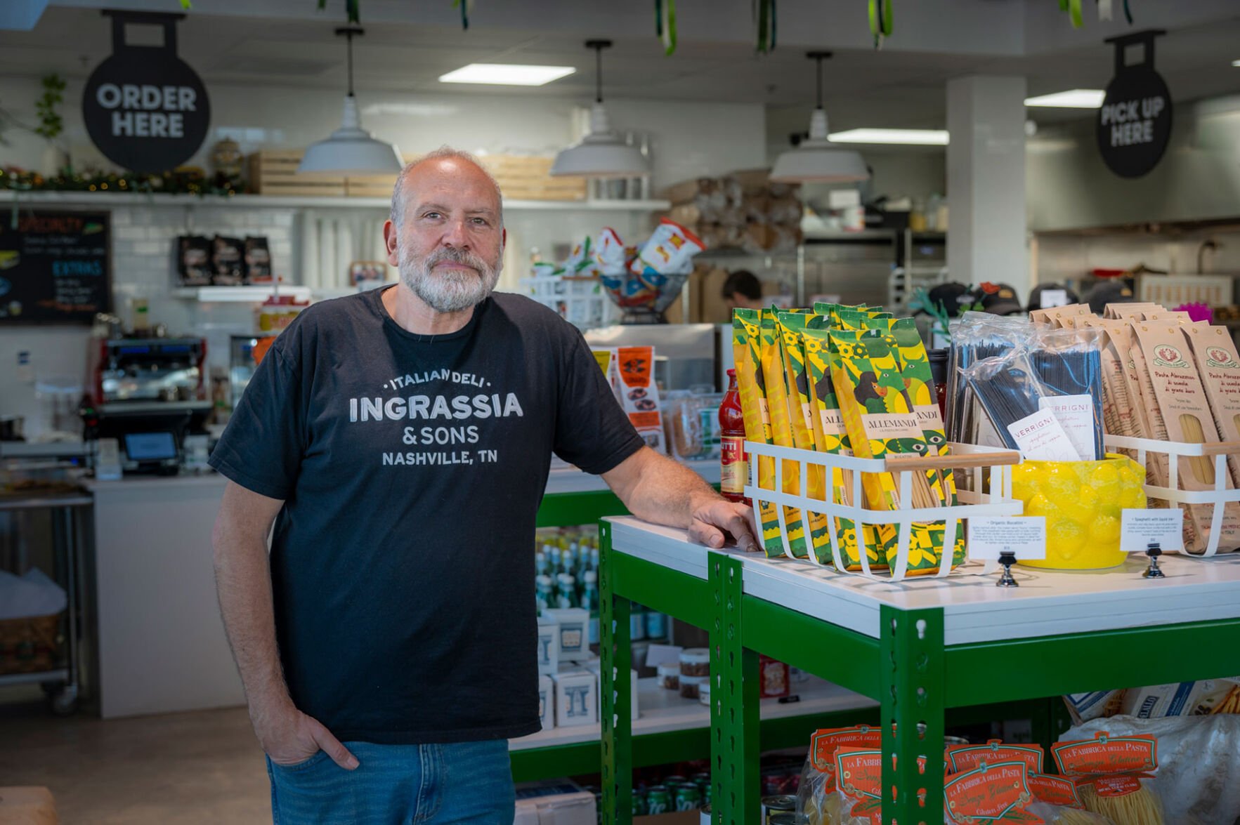 A man in a black t-shirt reading "Ingrassia & Sons" poses in the middle of a deli