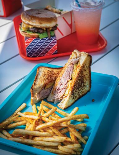 A ham sandwich with fries on a blue tray, with a burger on a red tray in the background