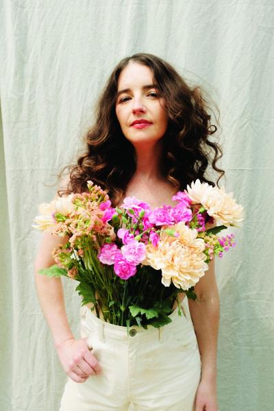 Color promo photo of the artist with a bouquet of flowers growing out of her trousers, worn as a kind of shirt