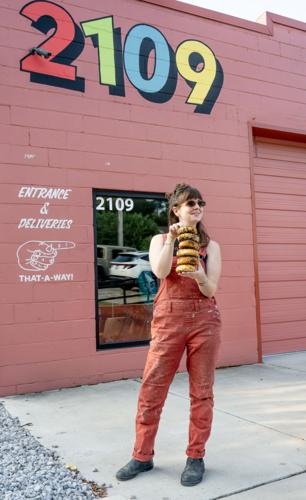 Kathleen Sharpe holding a stack of bagels in front of the All or Nothing Bagel storefront, a pink building with a multicolored number 2109 near the top of the facade