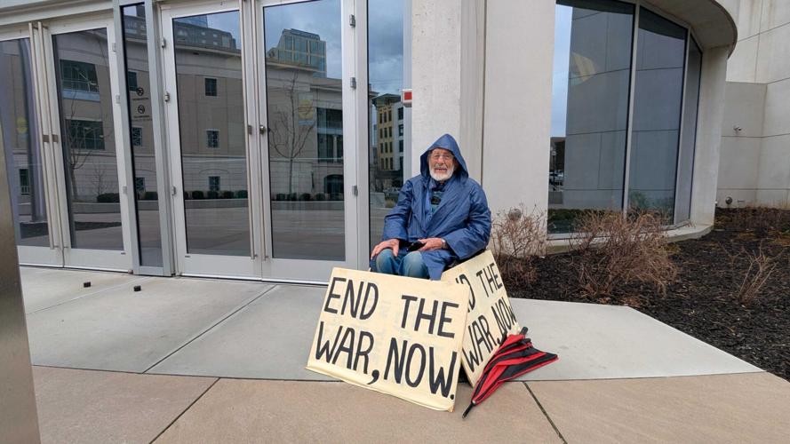 A man in a blue rain jacket sits outside a building with three signs all reading “END THE WAR, NOW.”