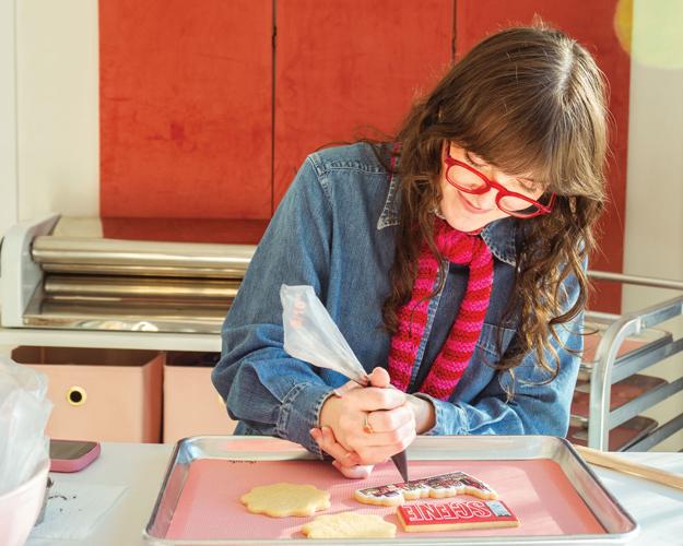 A woman in bright red glasses with a purple-and-red scarf pipes frosting onto a cookie