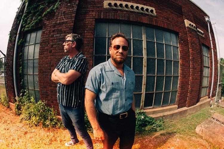 Color press photograph, shot with a slightly distorted fisheye lens, of The Black Keys’ Patrick Carney and Dan Auerbach standing outside a brick building showing signs of disuse.