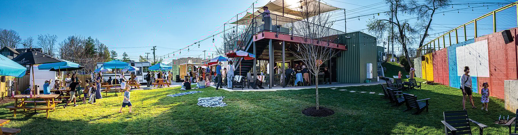 A panoramic shot of a grassy field with picnic benches, diners and food trucks near a boxy structure with a roof deck