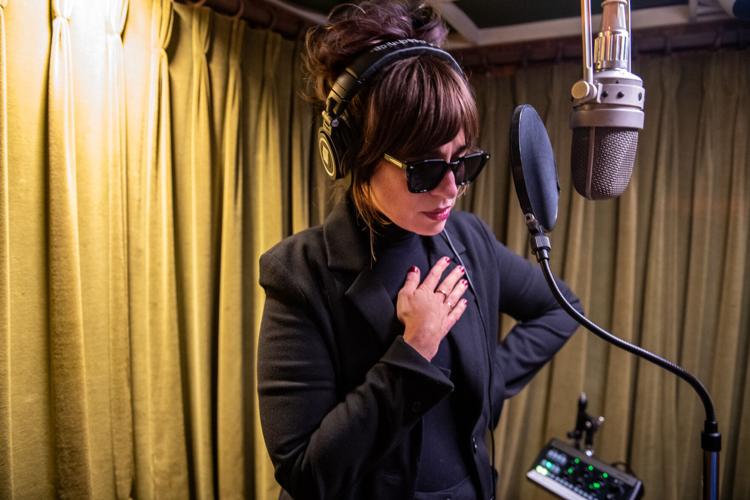 Color press photo of Nicole Atkins in the studio, dressed in black and wearing sunglasses, standing in front of a vocal microphone hung from above.