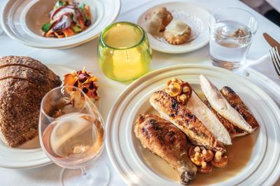 A plate of sliced chicken, including. drumstick, topped ith mushroom next to a glass of wine, a sliced loaf of bread and small bowl of salad