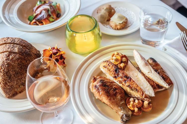 A plate of sliced chicken, including. drumstick, topped ith mushroom next to a glass of wine, a sliced loaf of bread and small bowl of salad