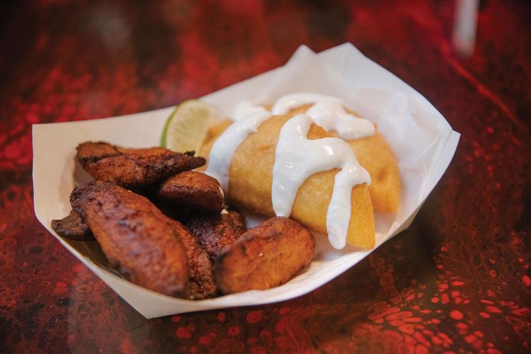 A small carboard tray with fried plantains and a pair of empanadas covered in cream
