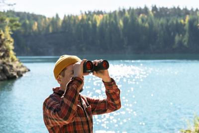 Young man, dressed in red plaid shirt, engaged in birdwatching with binoculars on the banks of calm blue mountain lake