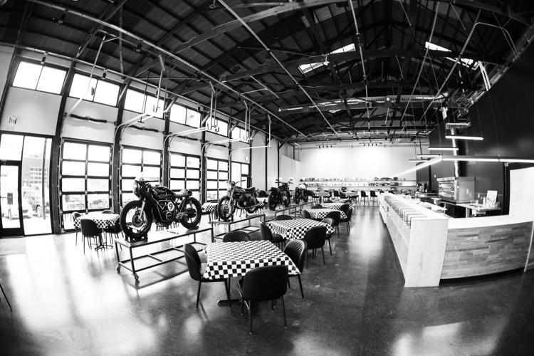A black-and-white photo of the interior of Moto Moda with a row of tables with checkered tablecloths adjacent to a row of four motorcycles