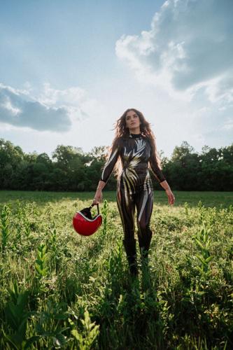 Color press photo of singer-songwriter Chloe Kimes for her ablum 'Daredevil,' dressed in a catsuit-type stage costume, holding a motorcycle helmet and walking through a field.
