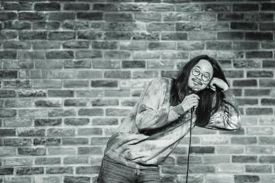 Black and white photo of comedian Sheng Wang, dressed in a sweater, performing in front of a brick wall.