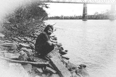 Black and white press photograph of songwriter John R. Miller sitting on the shore of a river with a bridge in the distance.