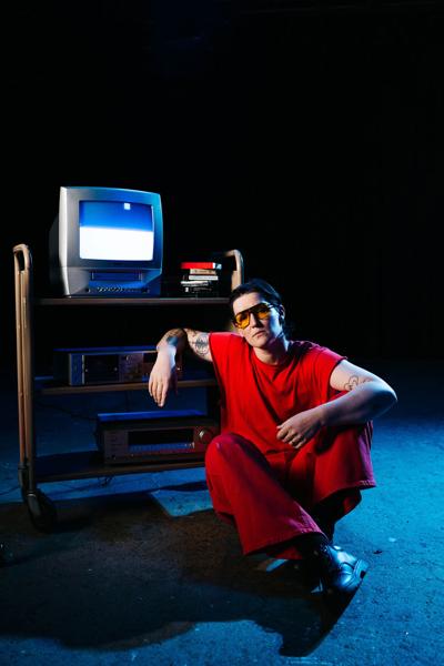 Artist photo of Semler, in which they wear a red jumpsuit and sunglasses in a dark room, seated on the floor with their arm on what looks like a school A/V cart