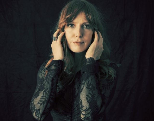 Color press photo of the artist looking into the camera, wearing a sheer black top and holding her hands by her face, standing against a black background.