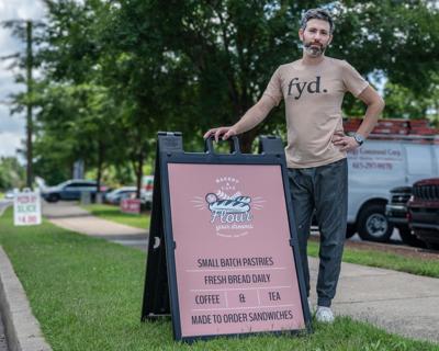 A man in a shirt reading “FYD.” stands next to a sandwich-board sign advertising the bakery Flour Your Dreams
