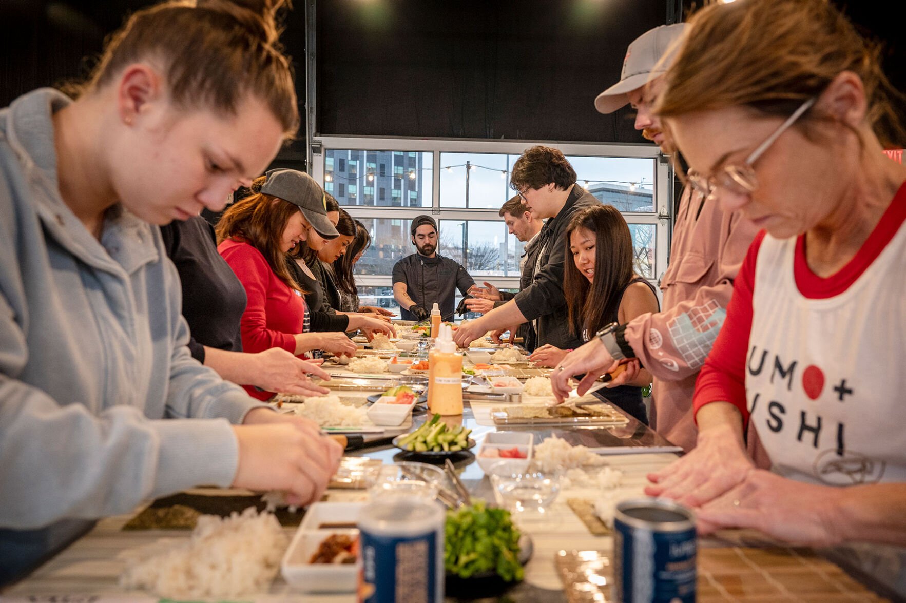 Eleven people huddle over a table rolling sushi, a chef at the end of the table instructing them.