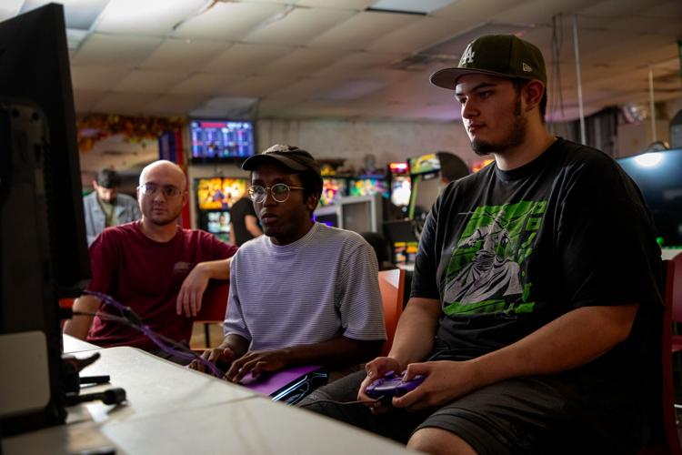 At Game Galaxy arcade, two players, one using a regular game controller and one with a portable arcade-style joystick controller, look at a TV while playing Street Fighter against each other. An onlooker leans in to view the match.
