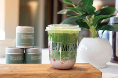 A matcha latte with strawberry syrup sits on a wooden board, a label reading "ceremony" stuck to the transparent cup