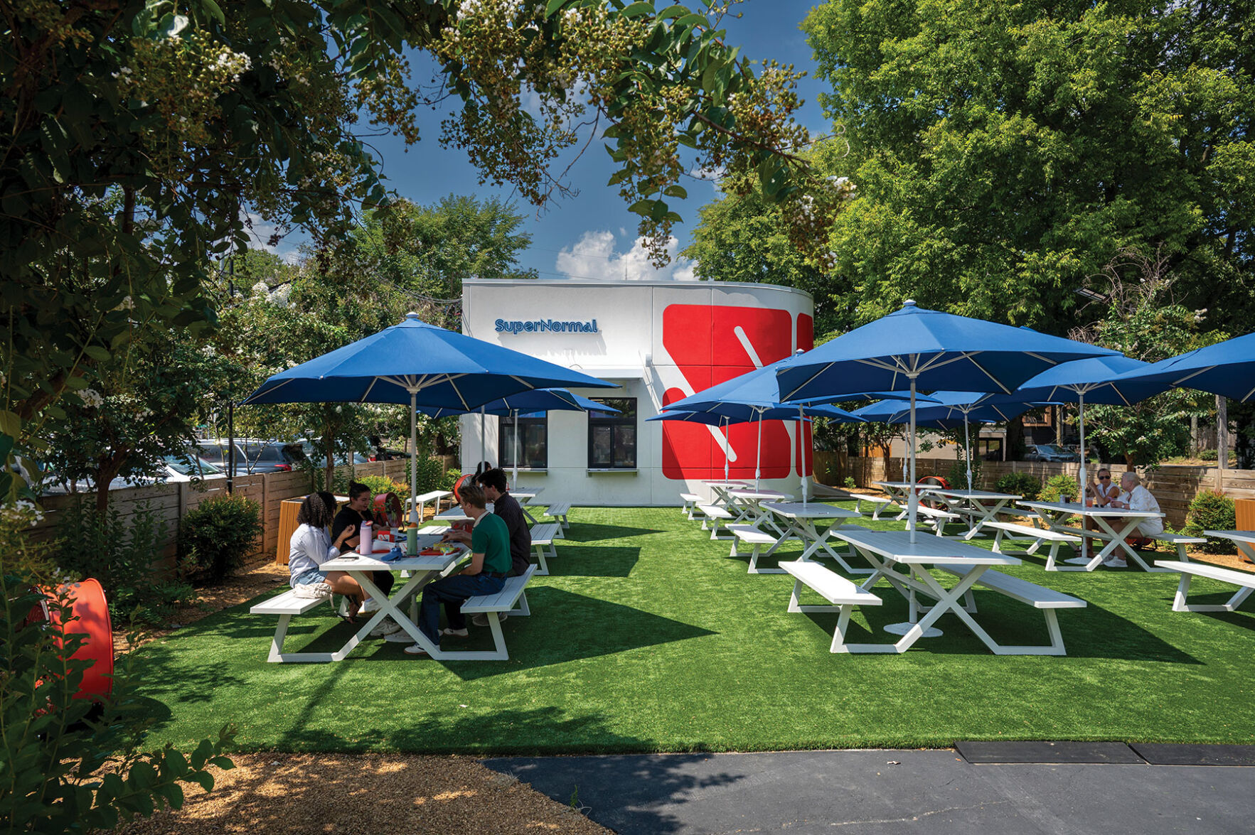 White park benches and blue umbrellas on the grass outside of SuperNormal's red and white building