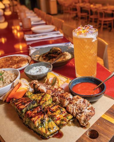 A wooden serving board holds filets of salmon and chicken skewers along with rice, sauces and an orange drink in a glass