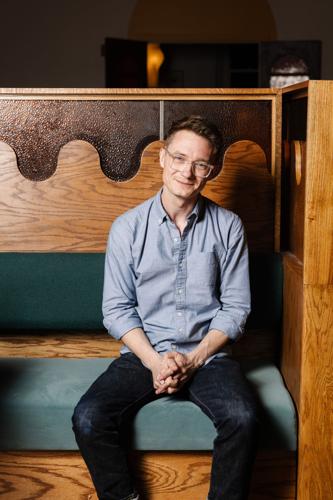 A smiling man with glasses in a blue shirt sits in a wooden booth