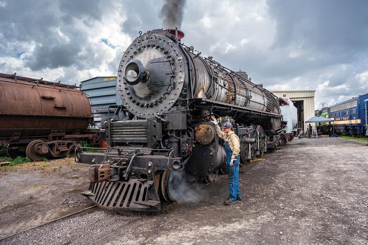 A man in overalls stands next to an old fashioned steam locomotive, smoke billowing from the top