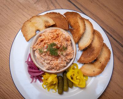 A bowl of fish dip on a plate surrounded by slices of toasted bread and pickled vegetables