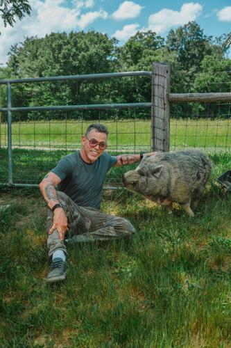 A man in sunglasses poses on the ground next to a hairy, tusked pig