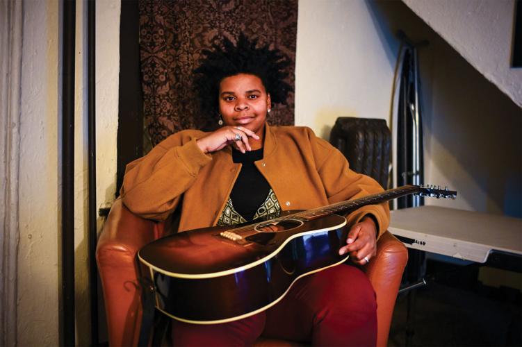 Joy Clark press photo artist seated in leather chair smiles and holds her guitar
