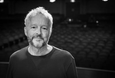 Black and white photograph of steel guitar player and music historian Pete Finney, standing onstage with an empty auditorium in the background.