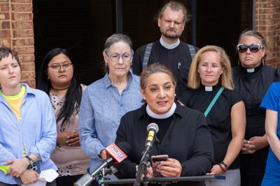 A woman in a black shirt and clergy’s white collar speaks at a lectern, a crowd behind her