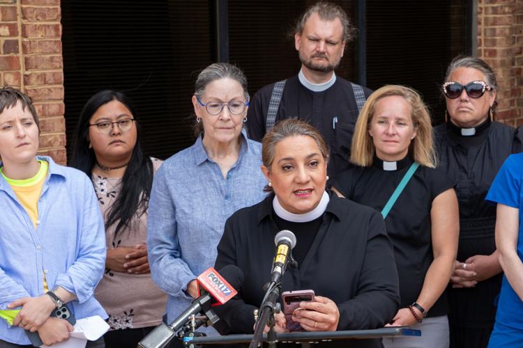 A woman in a black shirt and clergy’s white collar speaks at a lectern, a crowd behind her
