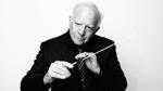 Black and white photo of Leonard Slatkin, dressed in black against a white background, holding his conductor's baton dramatically.