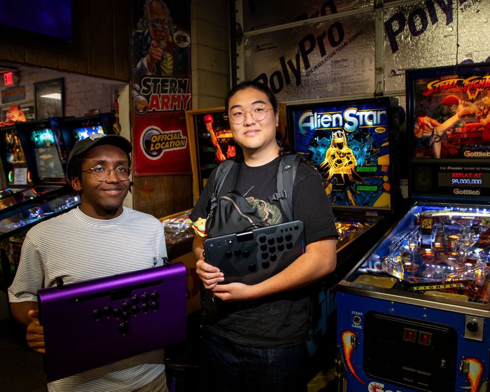 Two Street Fighter players pose in the pinball room at Game Galaxy next to a Street Fighter-themed pinball machine. They hold up their game controllers that mimic arcade joysticks and buttons.