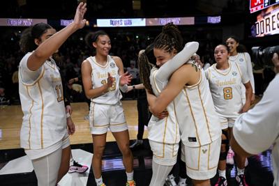 Two women's basketball players hug on the court while other teammates celebrate nearby. They wear white uniforms lined with gold.