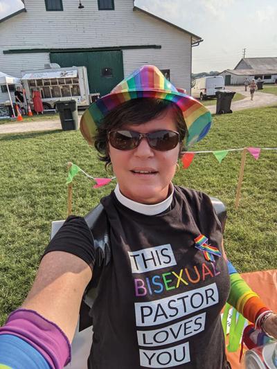 A woman with a pastor's collar wearing a rainbow colored hat and a shirt that reads "This Bisexual Pastor Loves You"