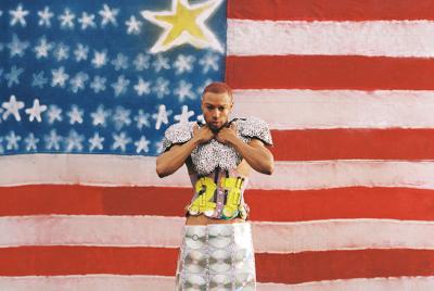 Color photo of the artist from the knees up, looking into the camera, standing in front of a large handmade American flag with stars spraypainted on. He’s dressed in a skirt covered in CDs, a pink corset with the number 27 painted across the front and football pads covered in drink can tabs arranged like silver sequins.