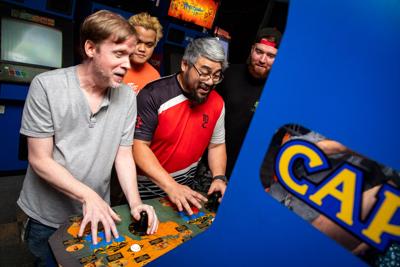 Two organizers of Game Galaxy's fighting game community face off on an arcade cabinet while two other organizer view the match.
