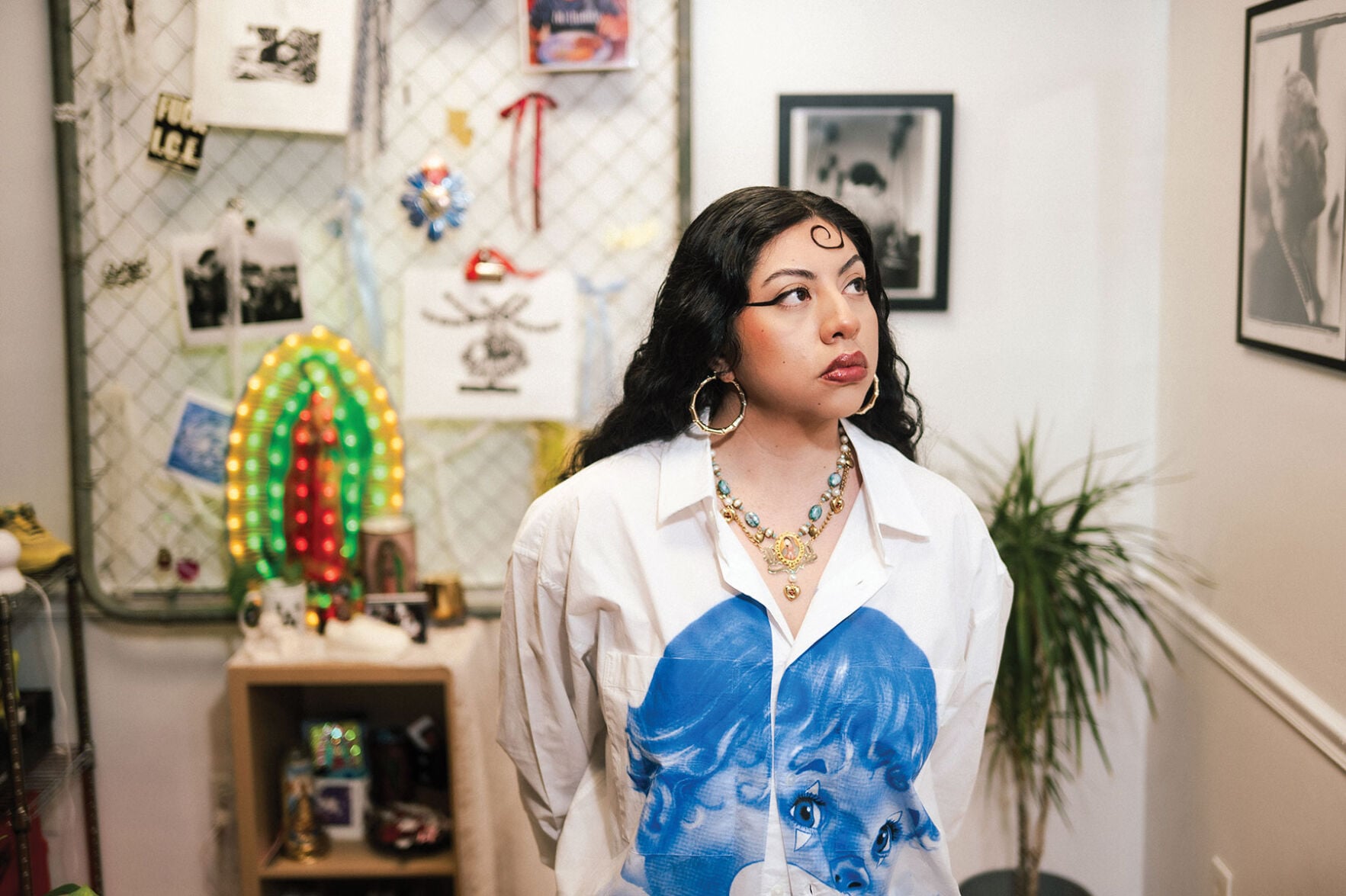 A woman with jewelry, dark hair and a white collared shirt with a blue printing of a childlish face on the front stares off to the right, a collection of trinkets and photos visible behind her.