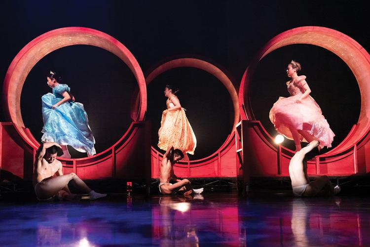 Three women in dresses run in gigantic hamster wheels, while three men sit in front dressed only in underwear