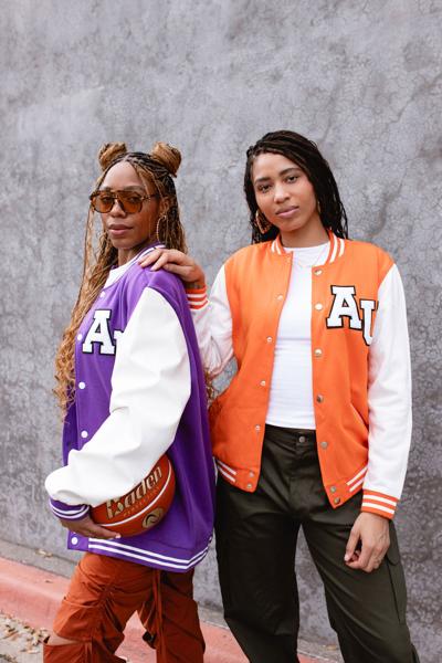 Two women stand next to each other. The one on the left wears sunglasses and a purple varsity jacket and holds a basketball. The one on the right wears an orange varsity basketball and rests a hand on the other woman's shoulder. Both have the letters "AU" on their jackets.
