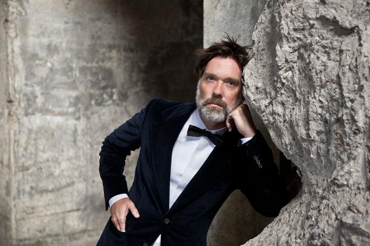 Rufus Wainwright press pic artist in tuxedo leans on a rock formation