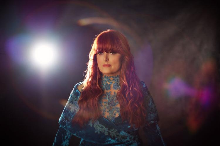 Color artist photo of Wynonna Judd, wearing a medium-blue dress with lace accents, standing against a gray backdrop with a slight prismatic shimmer. She looks into the camera as a bright light over her right shoulder creates a lens flare.