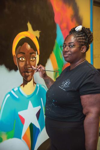 An artist paints a large  colorful mural of a girl with a large afro and blue t-shirt with a star on it.