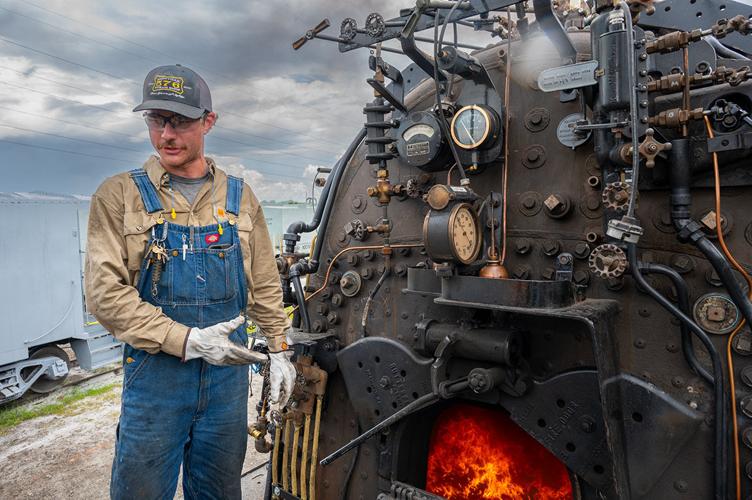 A an in a hat and overalls points to the boiler of a locomotive, flames glowing within