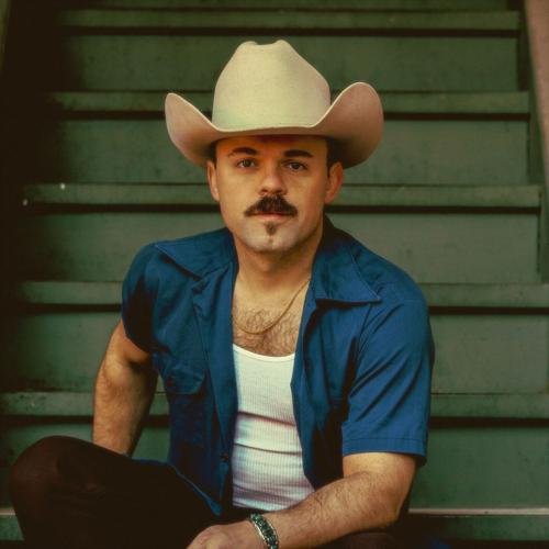 Sitting on green staircase, artist looks into the camera wearing a tan cowboy hat, a blue short-sleeve shirt open over a tank top and black jeans