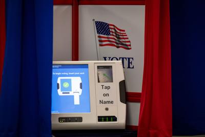 A voting machine with a screen displaying illustrated directions on how to insert a ballot. A sign reading "vote" with an American flag is displayed in the background.