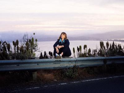 Color photo of the artist, taken near dusk or near dawn, sitting on a guardrail by the side of a road with a lake in a valley visible behind her