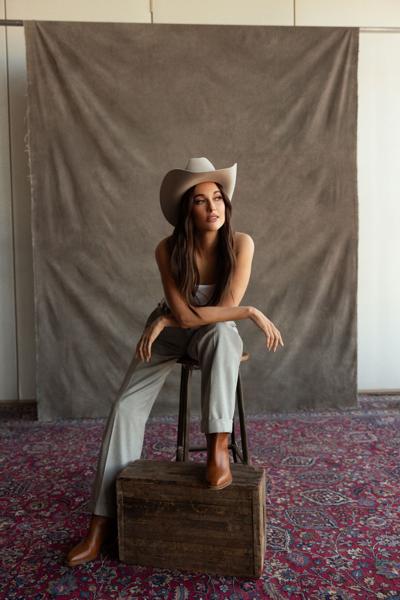 Color photo of the artist wearing a casual Western outfit including cowboy hat and boots, seated in a photo studio but with the studio infrastructure visible.